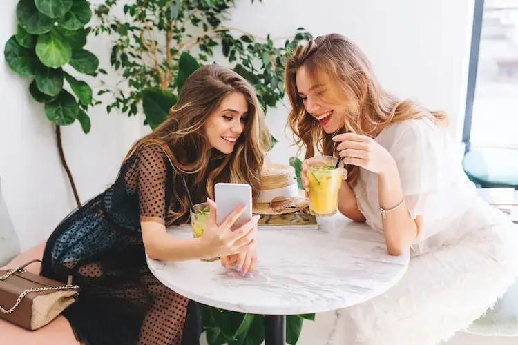 Two long-haired girls resting in cafe with modern interior and laughing. Indoor portrait of funny smiling ladies in trendy clothes making selfie while drink fruit cocktails.